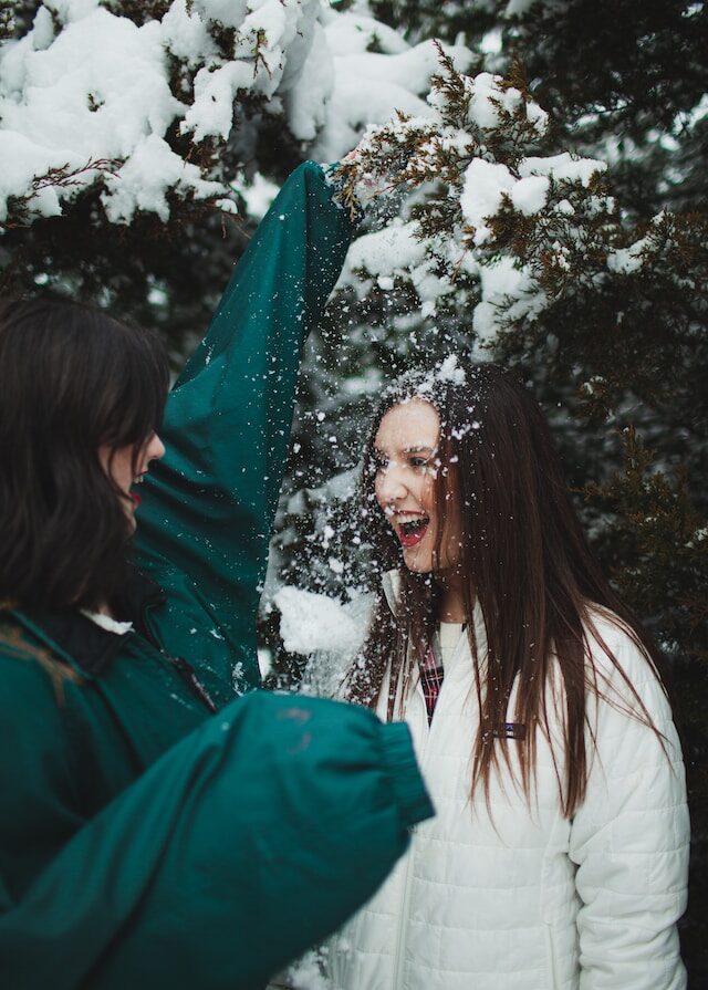 Nuori, tummatukkainen, vihreätakkinen karistelee lunta kuusesta toisen naisen päälle